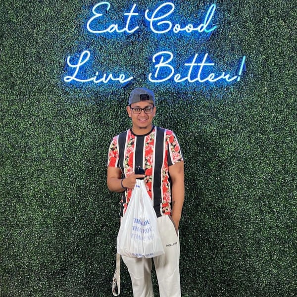Man holding a Supreme Empanadas takeout bag in front of a green wall with a neon sign.