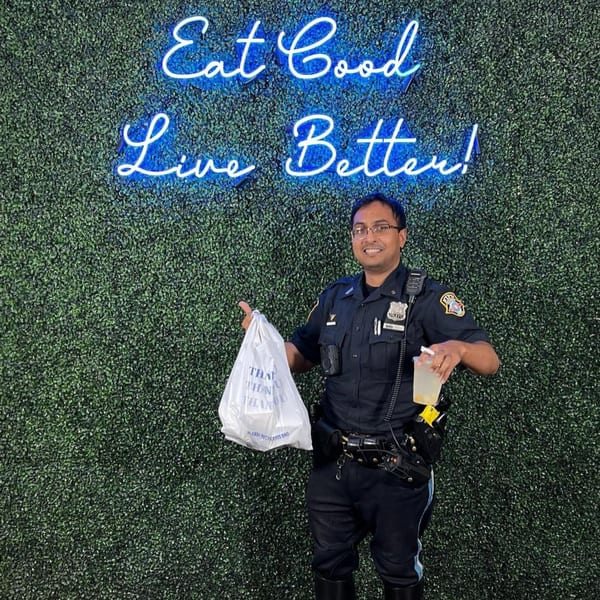 A police officer holding a Supreme Empanadas takeout bag and drink, standing before a neon sign.