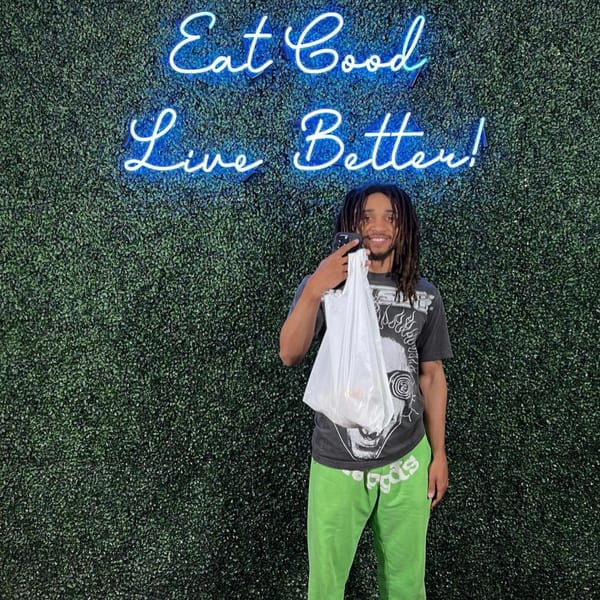 A man holding a white bag stands in front of a green wall with a blue neon sign.