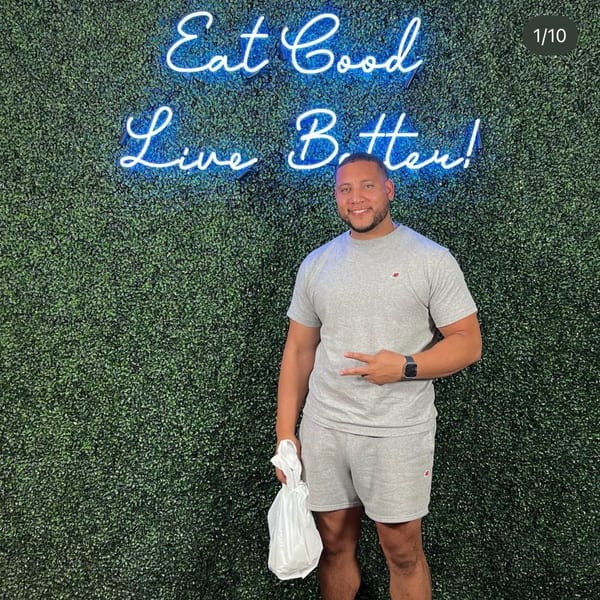Man holding a takeout bag, standing in front of a green wall with a neon "Eat Good Live Better!" sign.