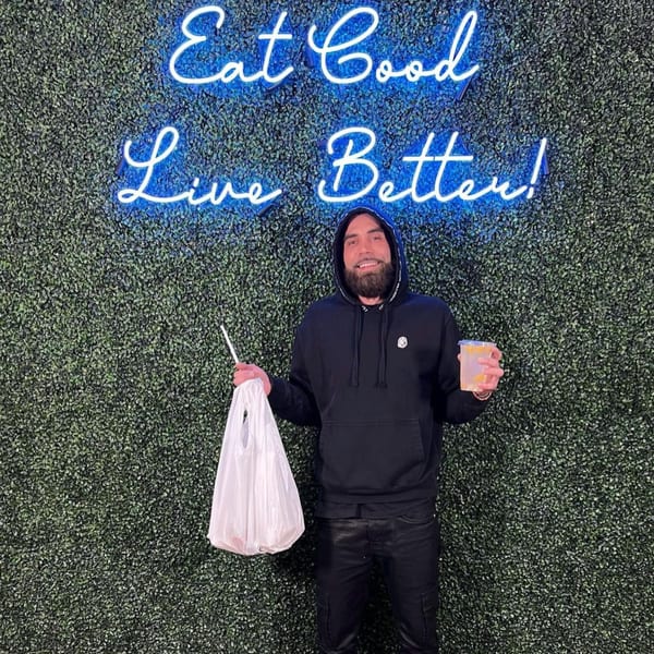 Man holding a Supreme Empanadas takeout bag and drink in front of a neon sign.